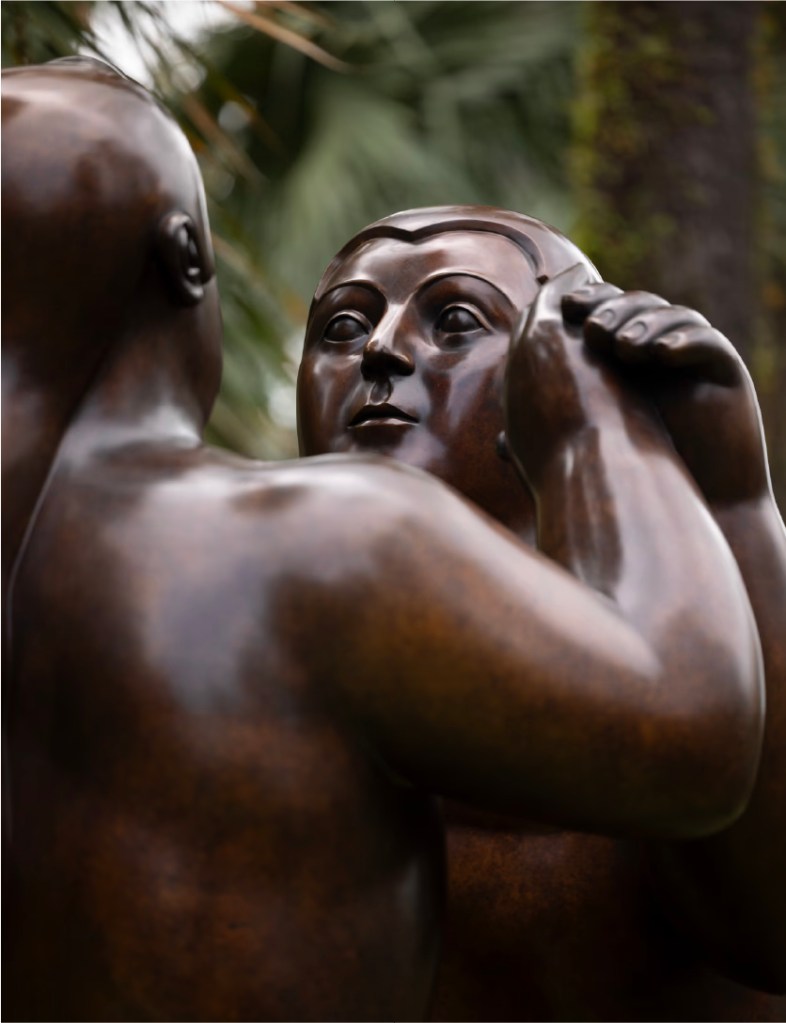 A monumental bronze sculpture of a voluminous dancing couple standing on a stone pedestal amidst the lush greenery of Gardens by the Bay.