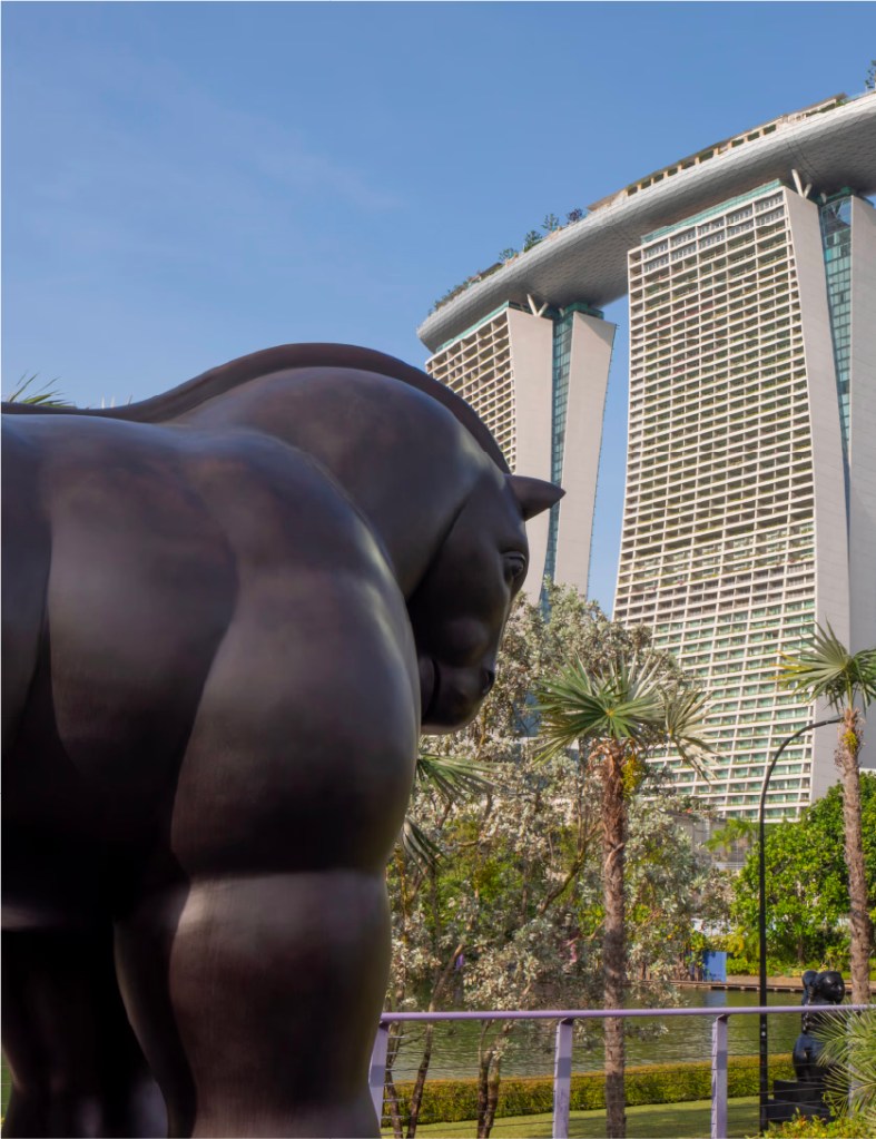 A monumental bronze sculpture of a voluminous horse standing on a stone pedestal amidst the lush greenery of Gardens by the Bay with a backdrop of Marina Bay Sands